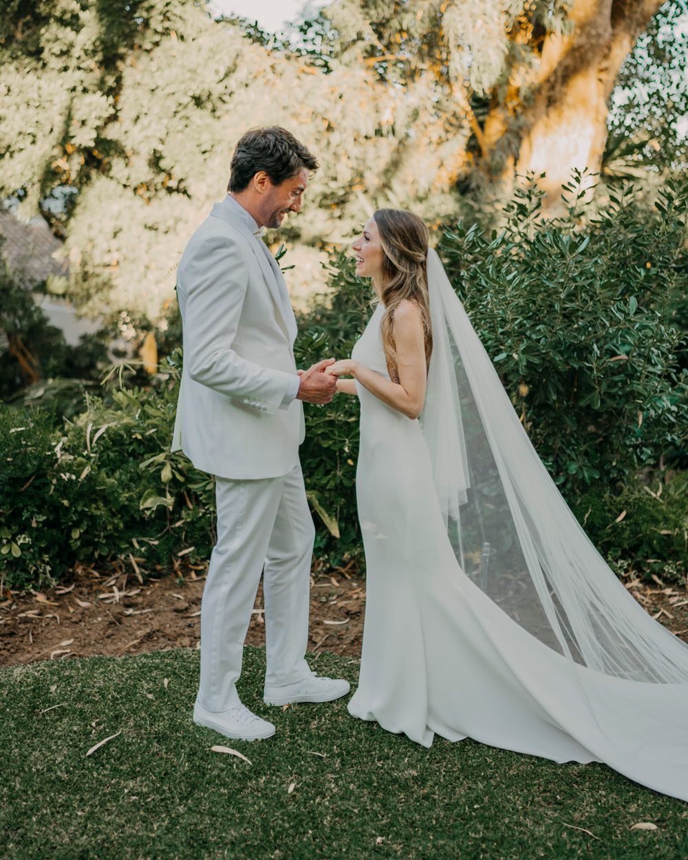 A couple in light-colored wedding attire holding hands in an outdoor garden setting with a long flowing veil.