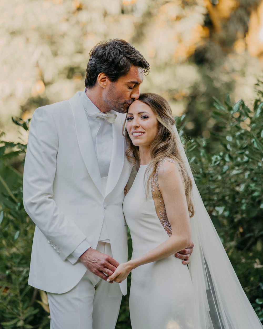 Newlywed couple in white attire embrace during sunset outdoor wedding photoshoot among greenery.