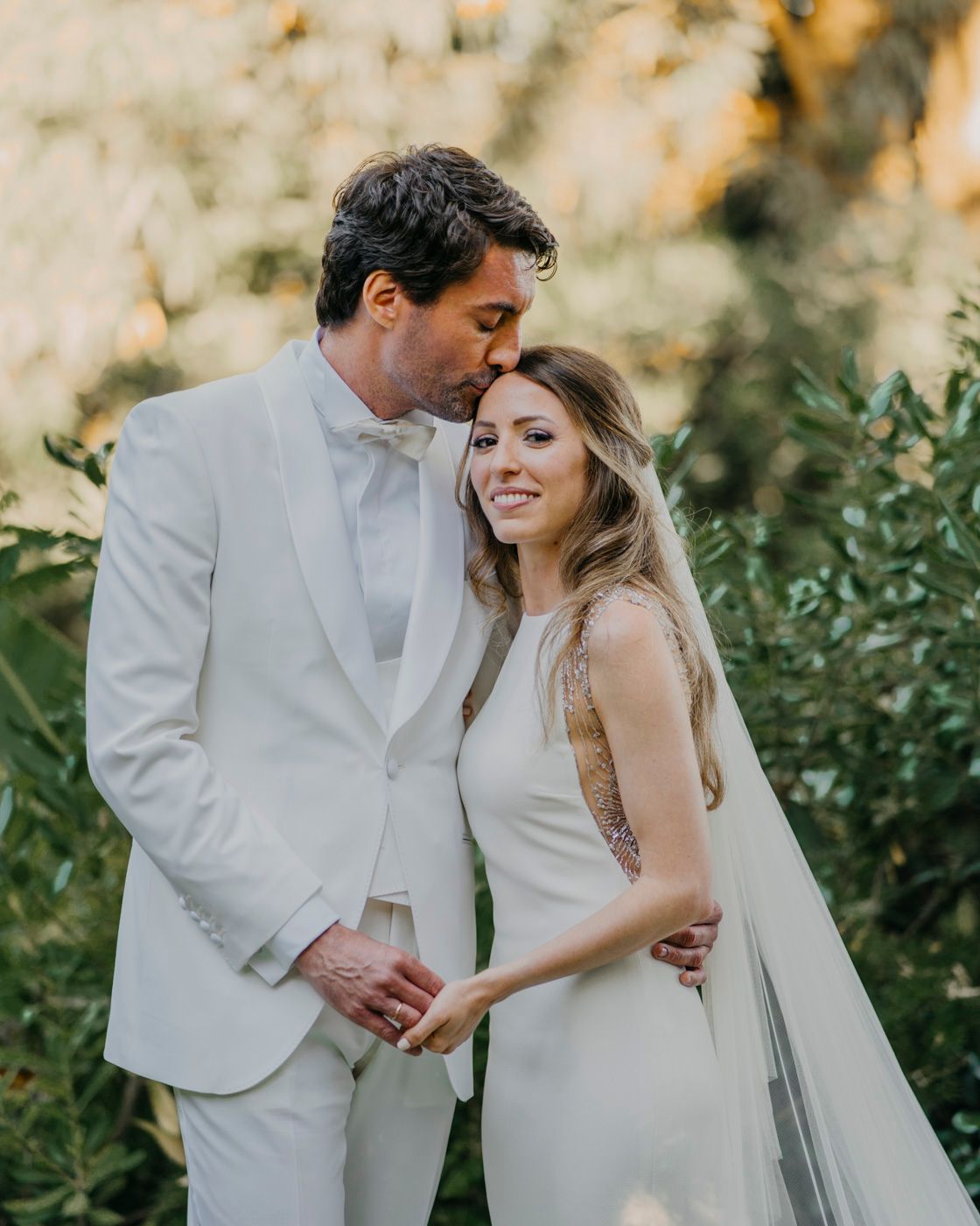 Newlywed couple in white attire embrace during sunset outdoor wedding photoshoot among greenery.
