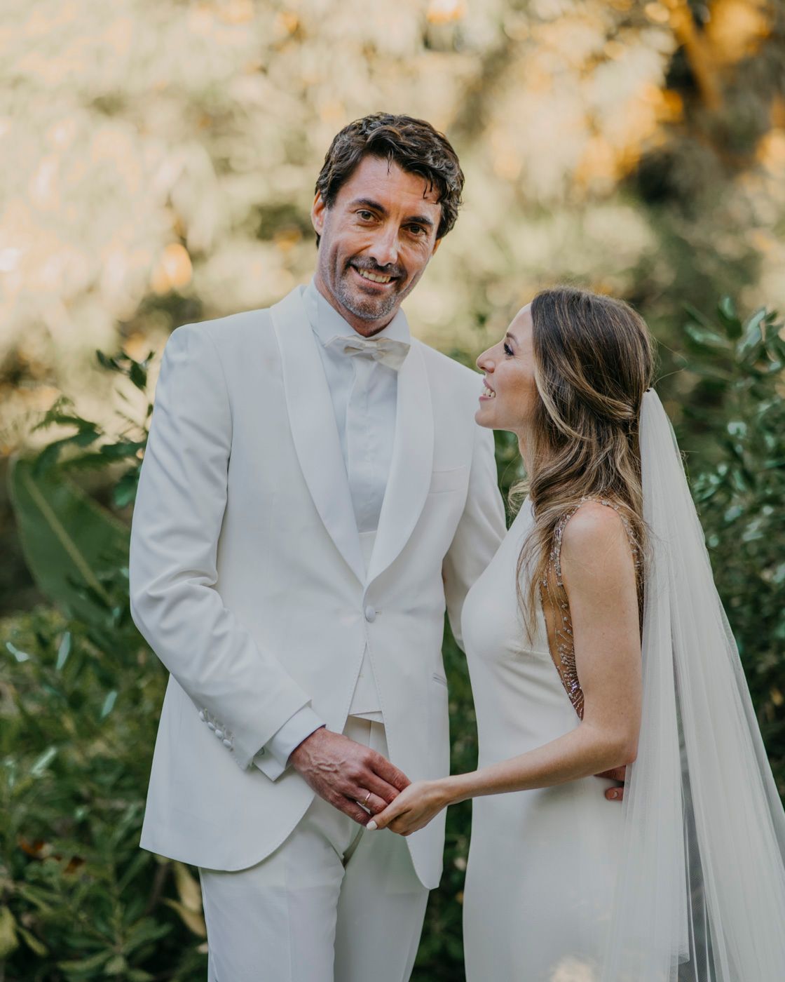 A bride and groom share a romantic moment in their white wedding attire against a sunlit garden backdrop.