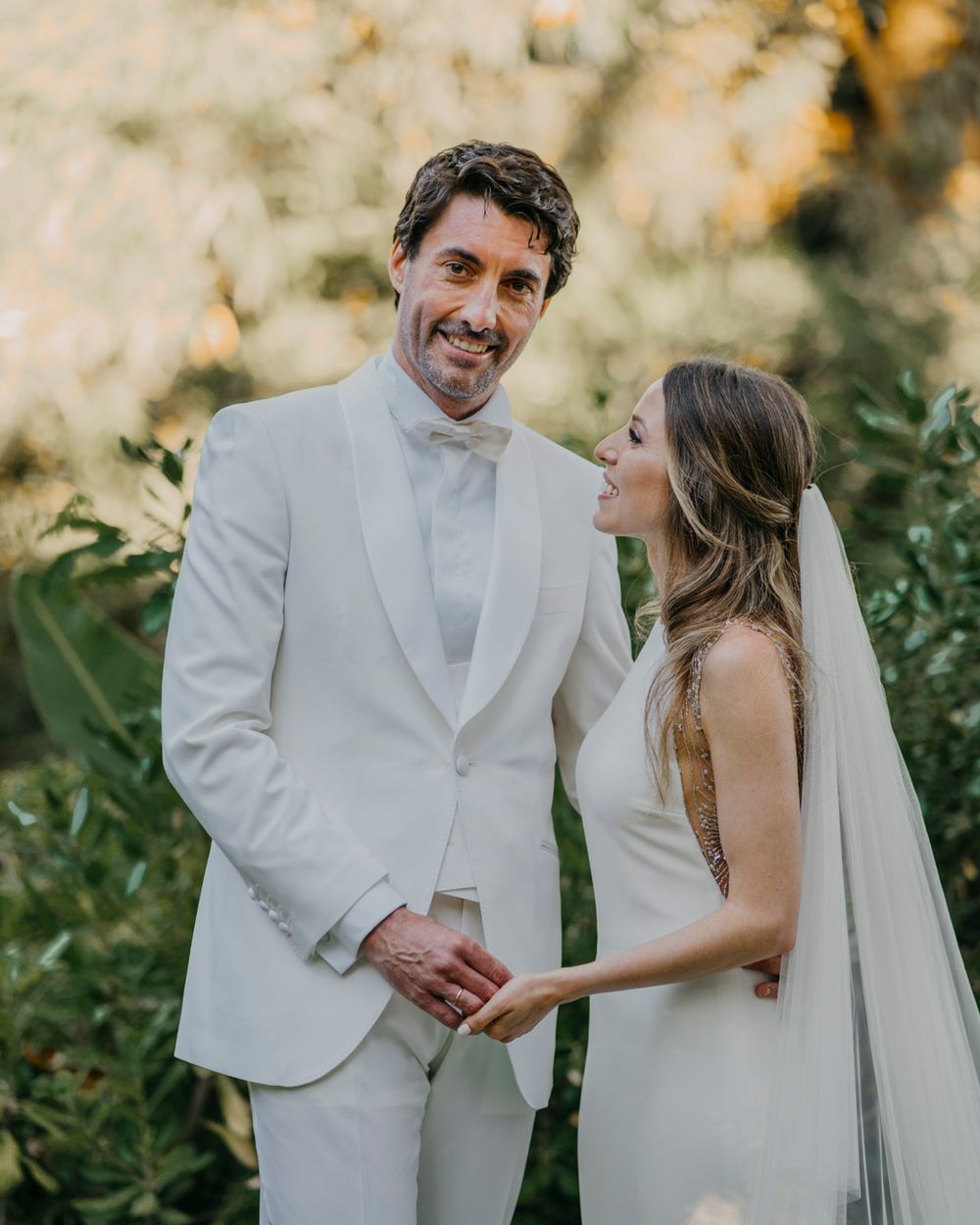A bride and groom share a romantic moment in their white wedding attire against a sunlit garden backdrop.