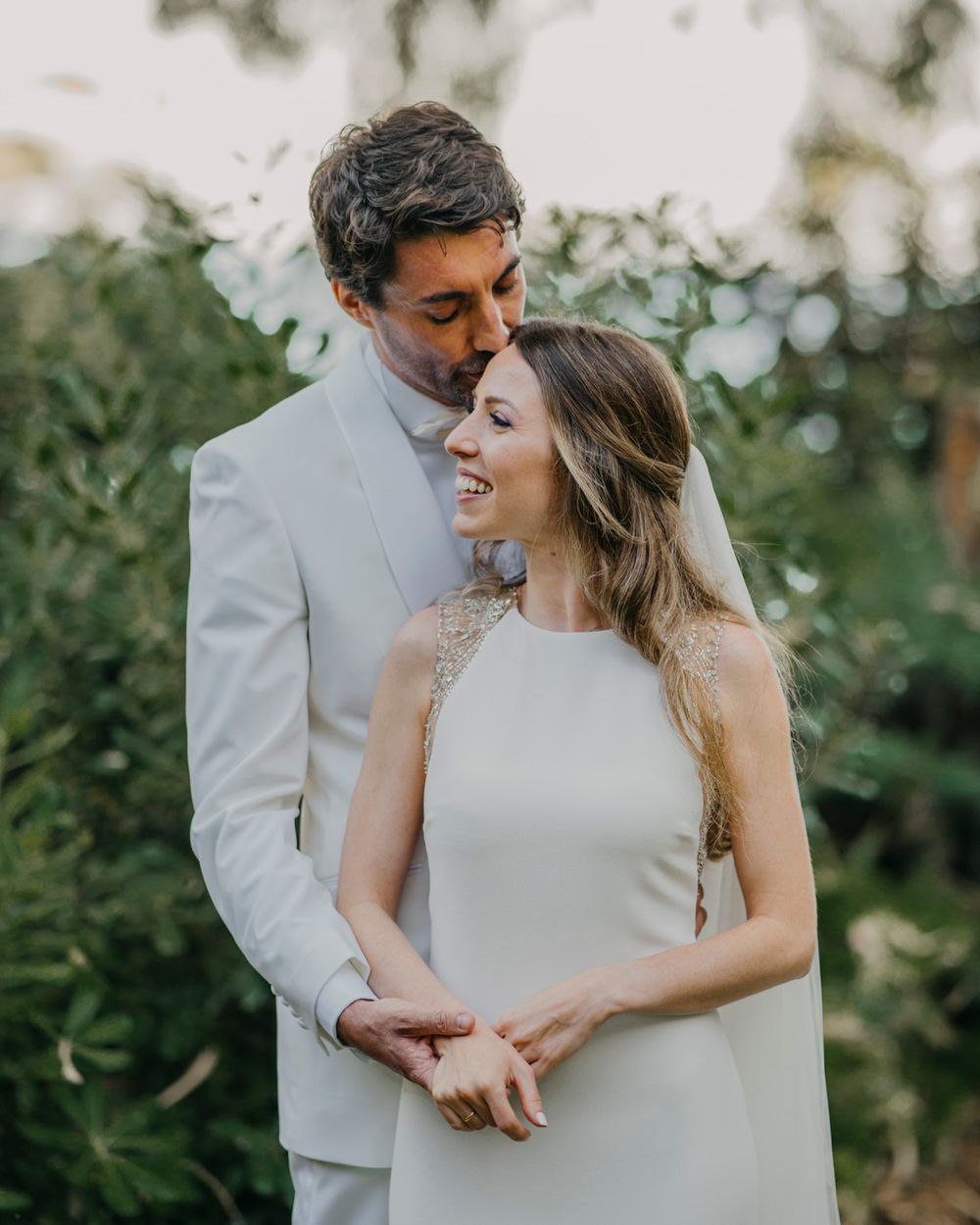A couple shares an intimate moment in a garden setting, with the bride in a sleek white dress and groom in light gray suit.