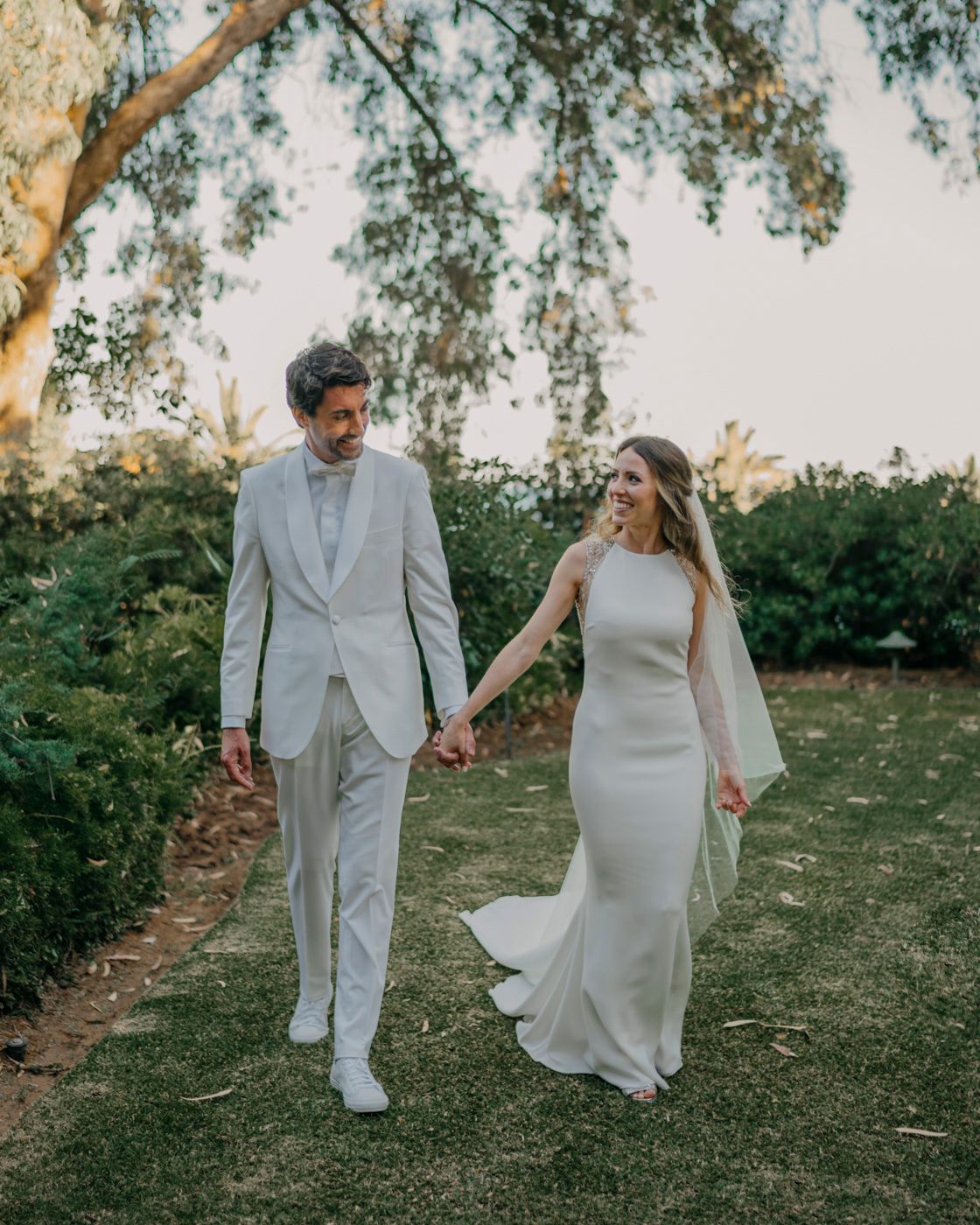 A couple in light-colored wedding attire walk hand in hand along a garden path under a tree at sunset.