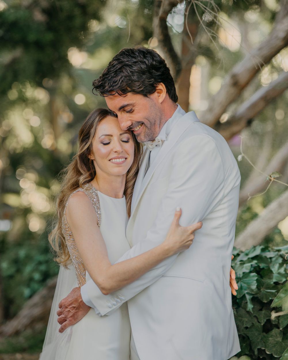 A tender intimate wedding moment between a couple in white attire embracing outdoors with soft natural lighting.
