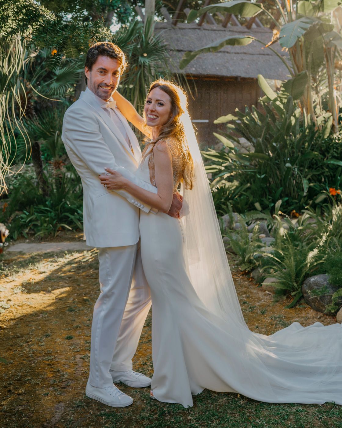 A couple shares a romantic moment in their white wedding attire surrounded by lush tropical garden foliage at sunset.