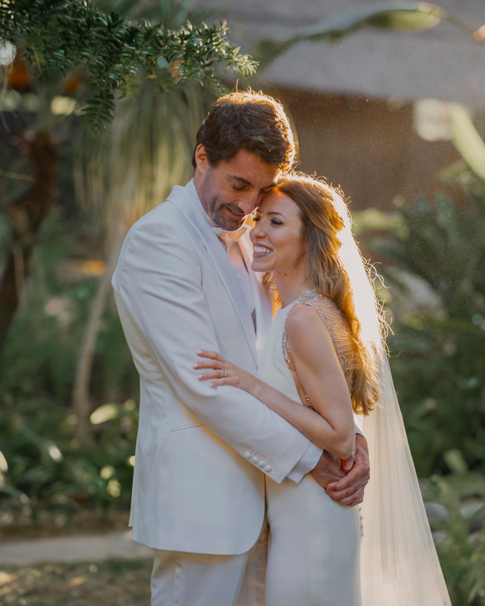 Romantic pair in white outfits share a tender embrace during a golden hour wedding ceremony in a garden setting.
