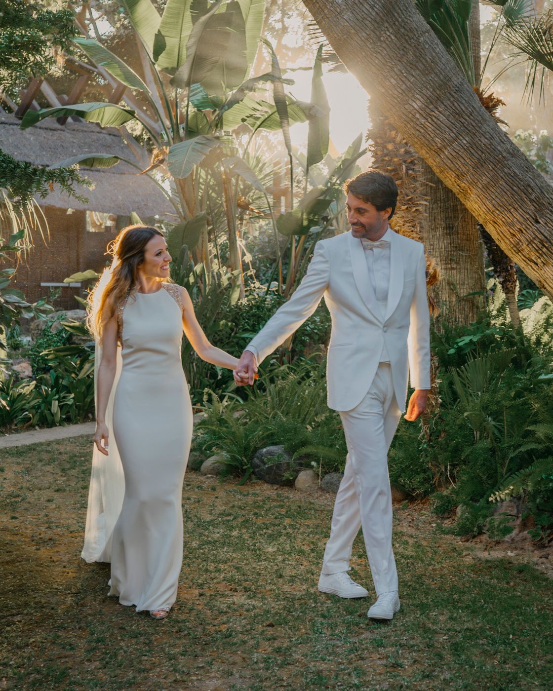 A couple in elegant white attire walk hand in hand through a lush tropical garden during a golden hour photoshoot.