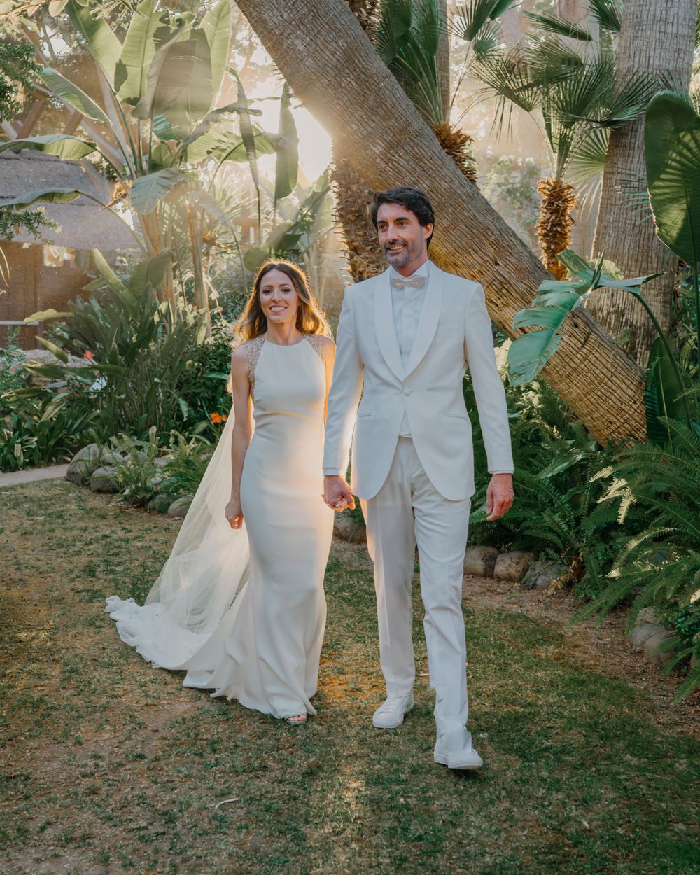 Two people in wedding attire walk hand in hand through a lush tropical garden setting with palm trees.
