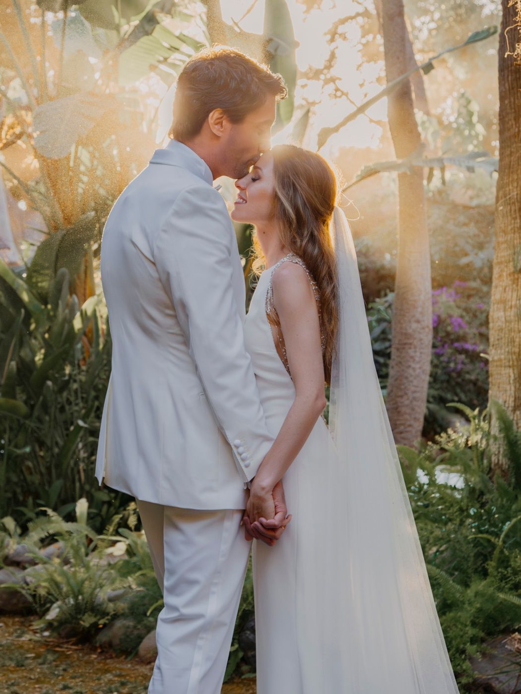 A romantic wedding couple shares an intimate moment in a sun-dappled garden setting with lush greenery and palm trees.