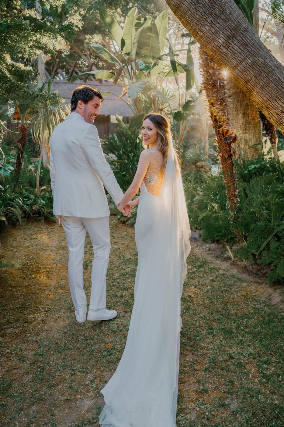 A couple in elegant white wedding attire holds hands in a lush garden setting with soft sunlight filtering through the trees.