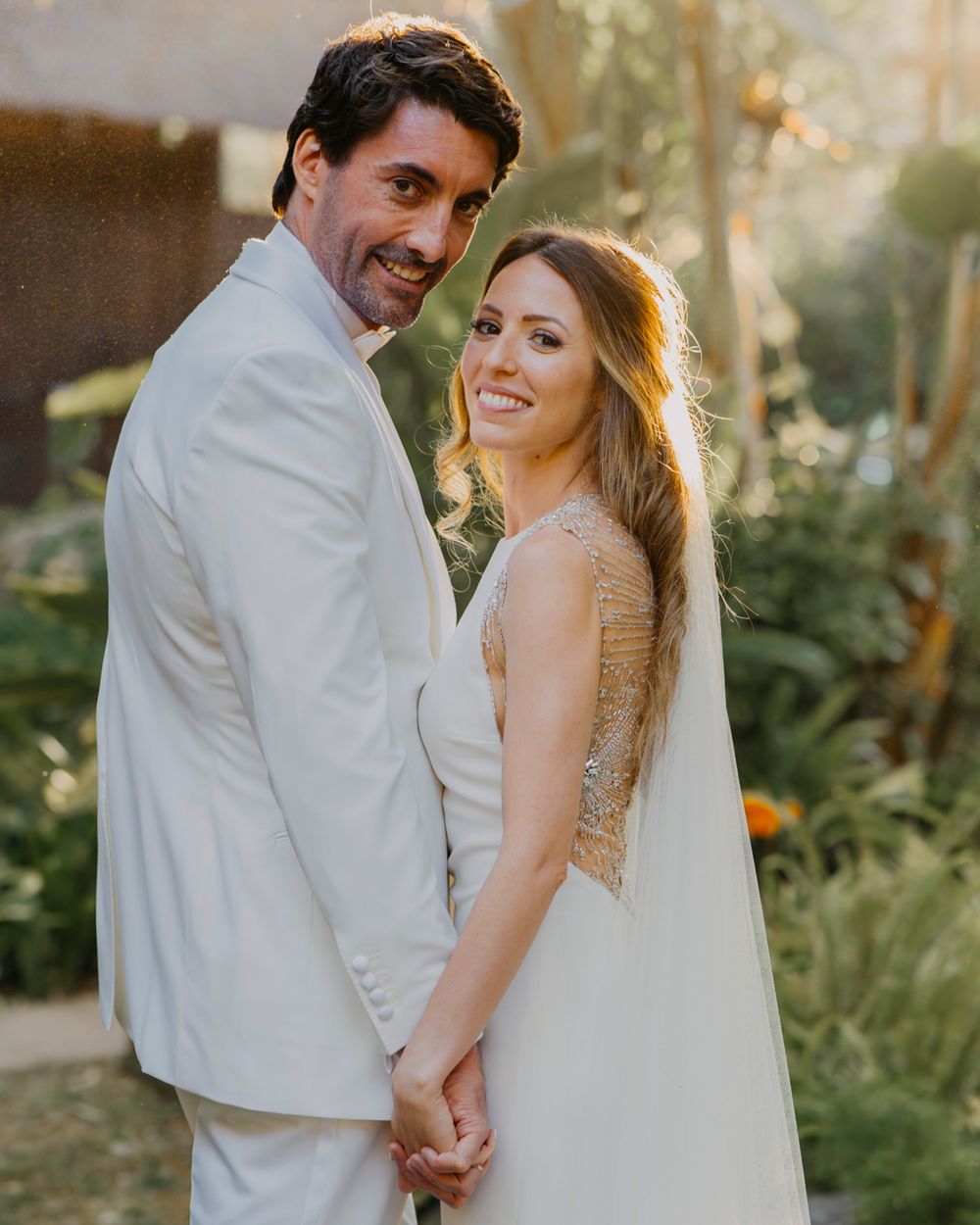 A couple in white wedding attire poses together in a garden setting during golden hour lighting.