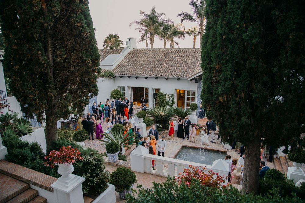 Elegant outdoor venue with thatched roof building, pool, and guests mingling in a lush garden setting.