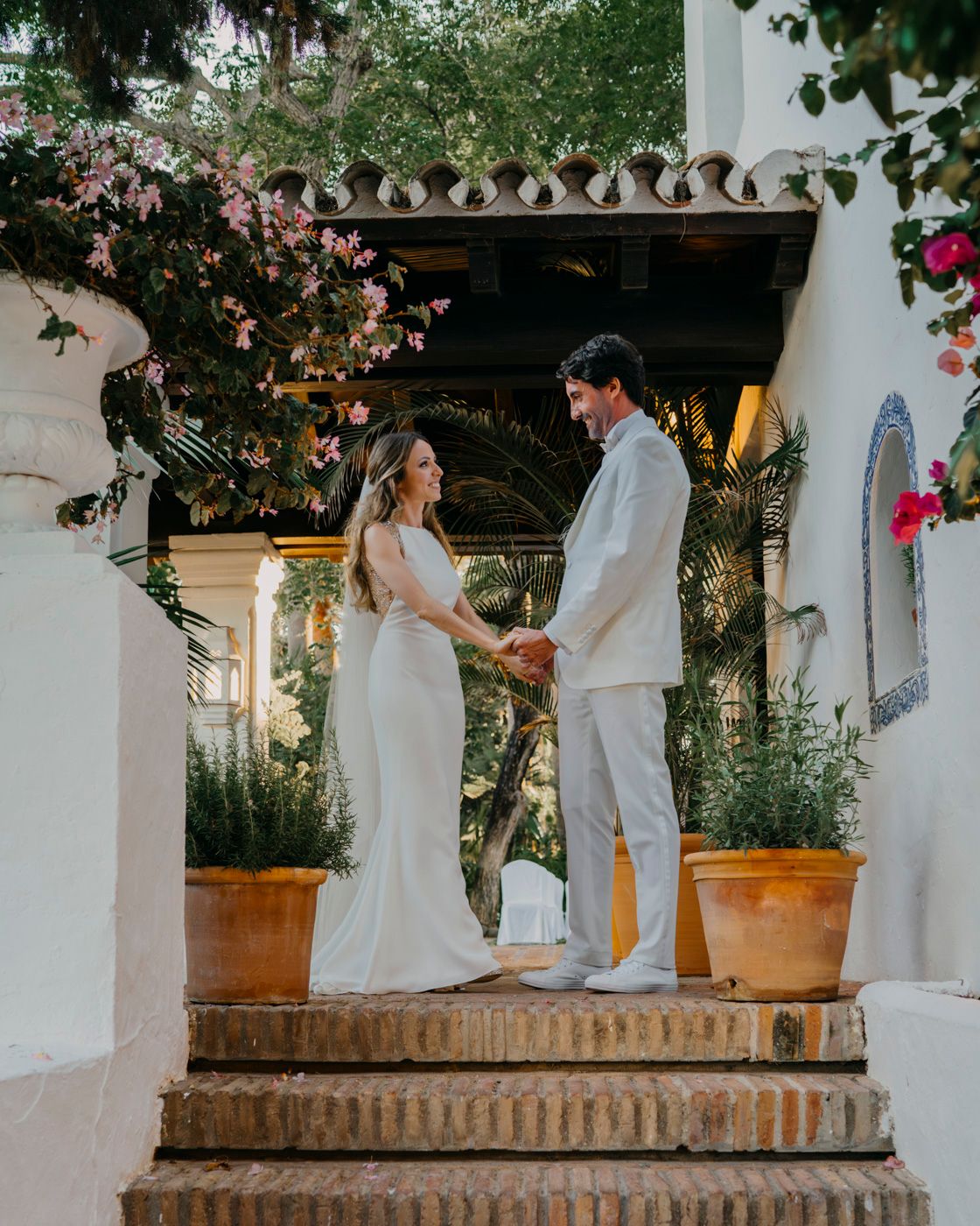 A couple in white wedding attire stands on stone steps at a Spanish villa surrounded by pink flowers and greenery.