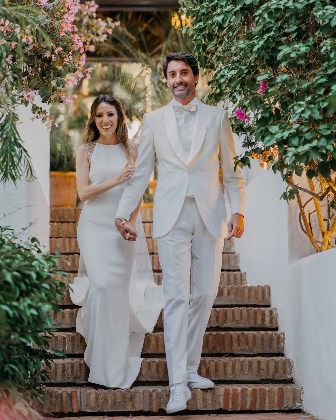 Wedding couple descends outdoor staircase surrounded by pink flowers and greenery in an elegant garden setting.