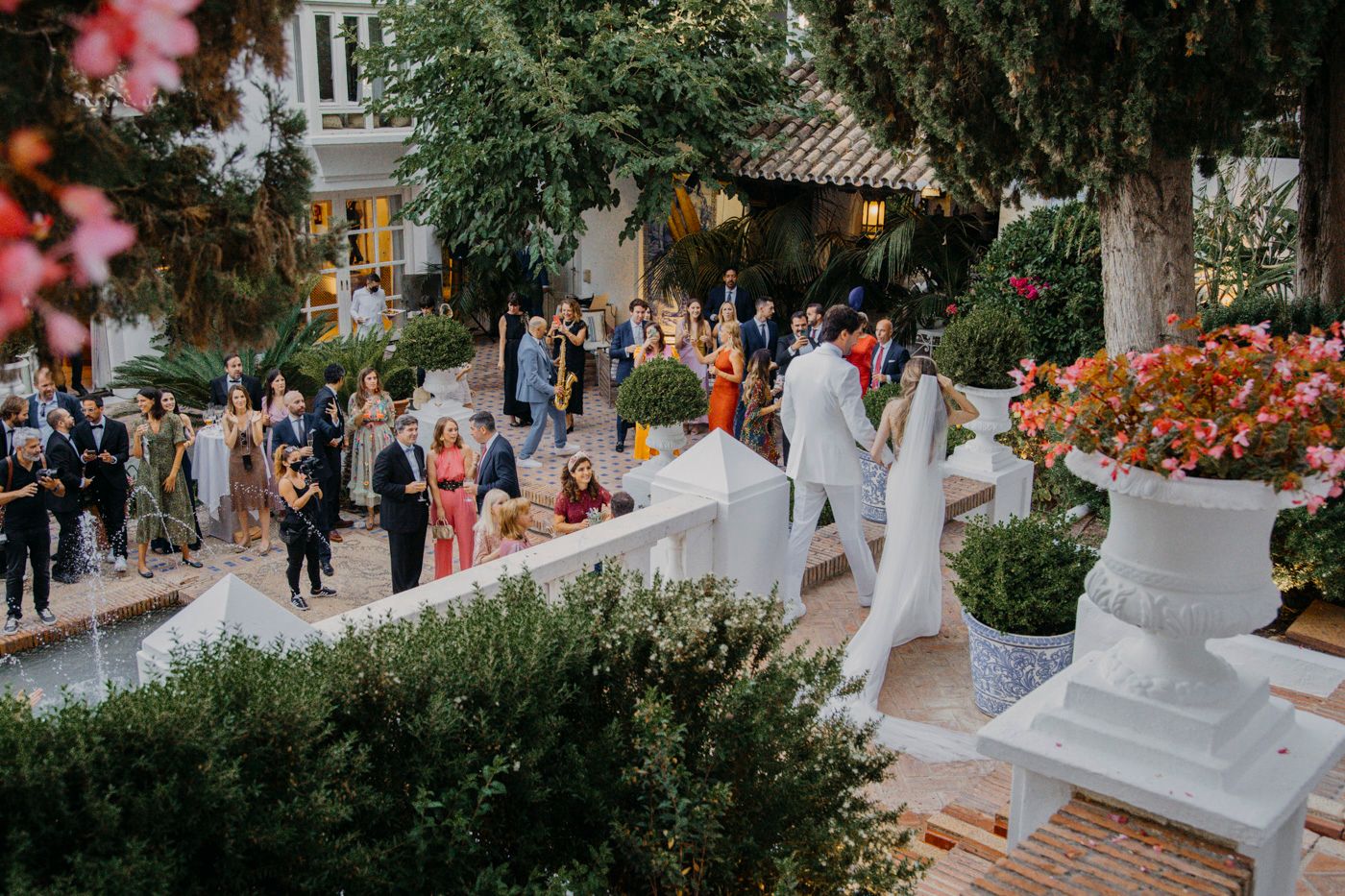 Outdoor wedding ceremony with guests gathered in a garden decorated with elegant white urns and colorful flowers.