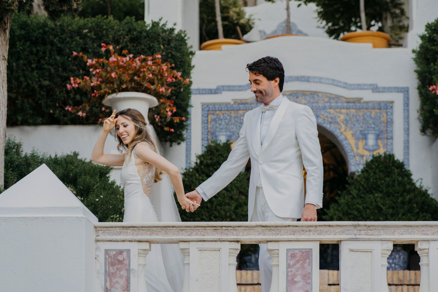 Newlyweds in white attire hold hands while walking outside a Mediterranean-style building with decorative tiles.