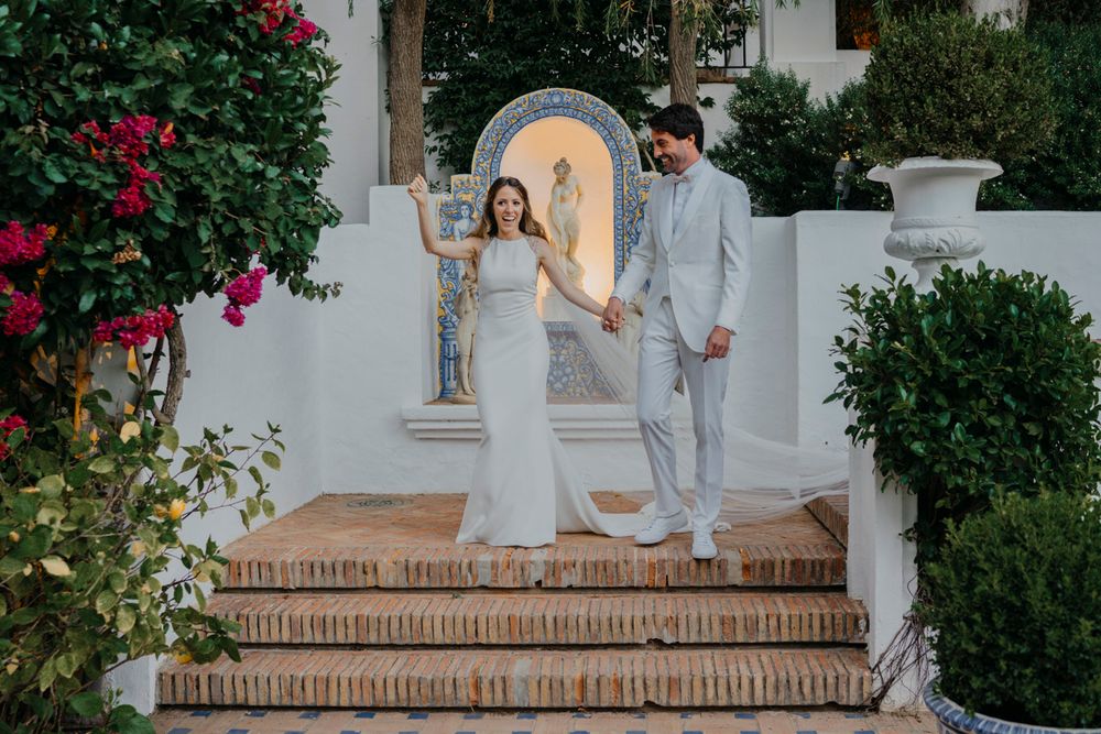 Wedding couple descending ornate Spanish-style stairs with white walls and blooming bougainvillea.