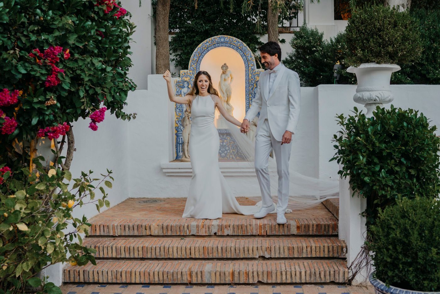 Wedding couple descending ornate Spanish-style stairs with white walls and blooming bougainvillea.