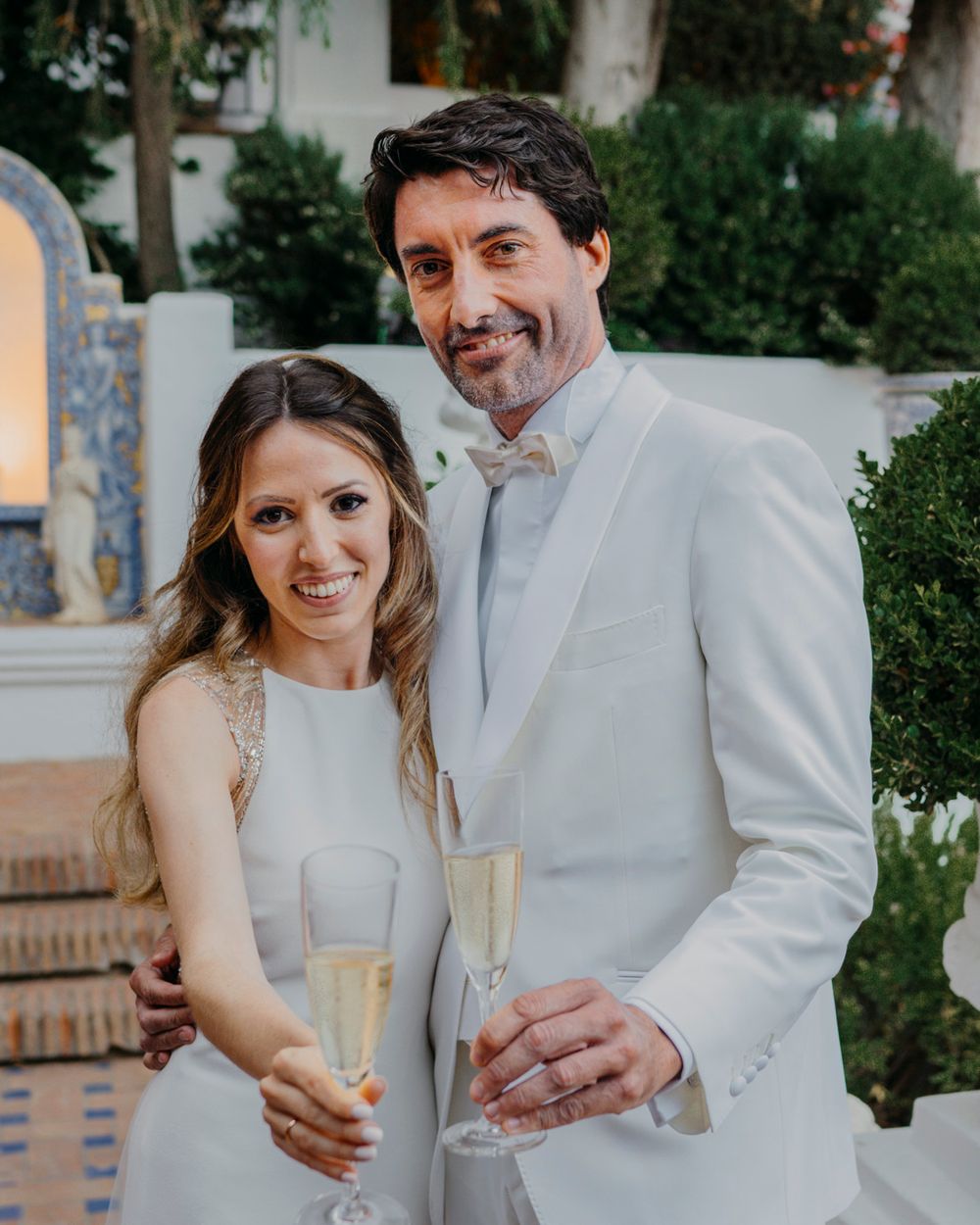 Couple in formal attire holding champagne glasses on Mediterranean-style terrace with arched alcove.
