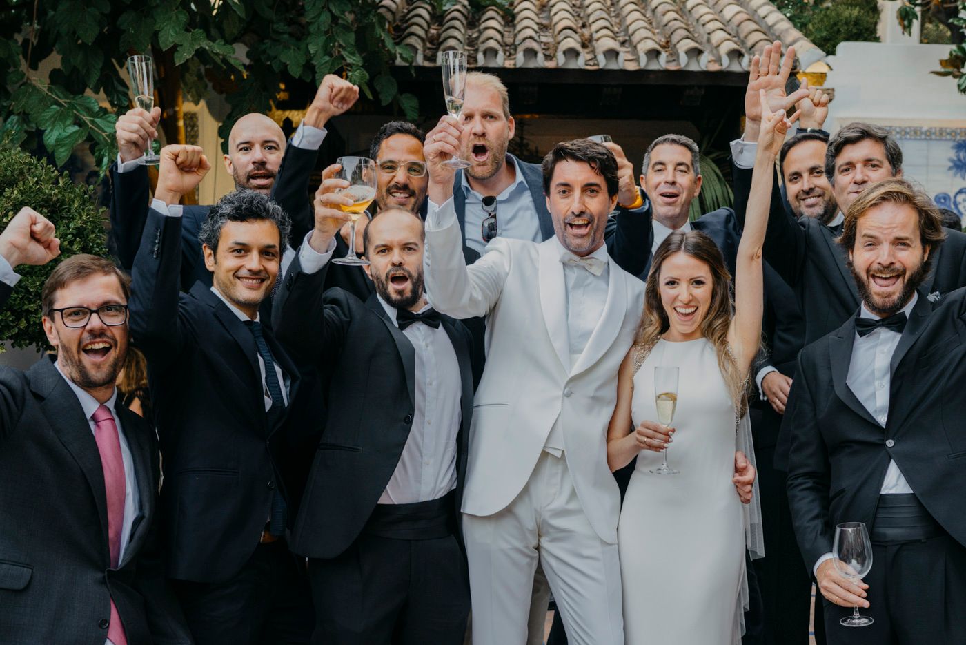 A celebratory group gathers together outdoors wearing formal attire with the couple in light colored wedding outfits.