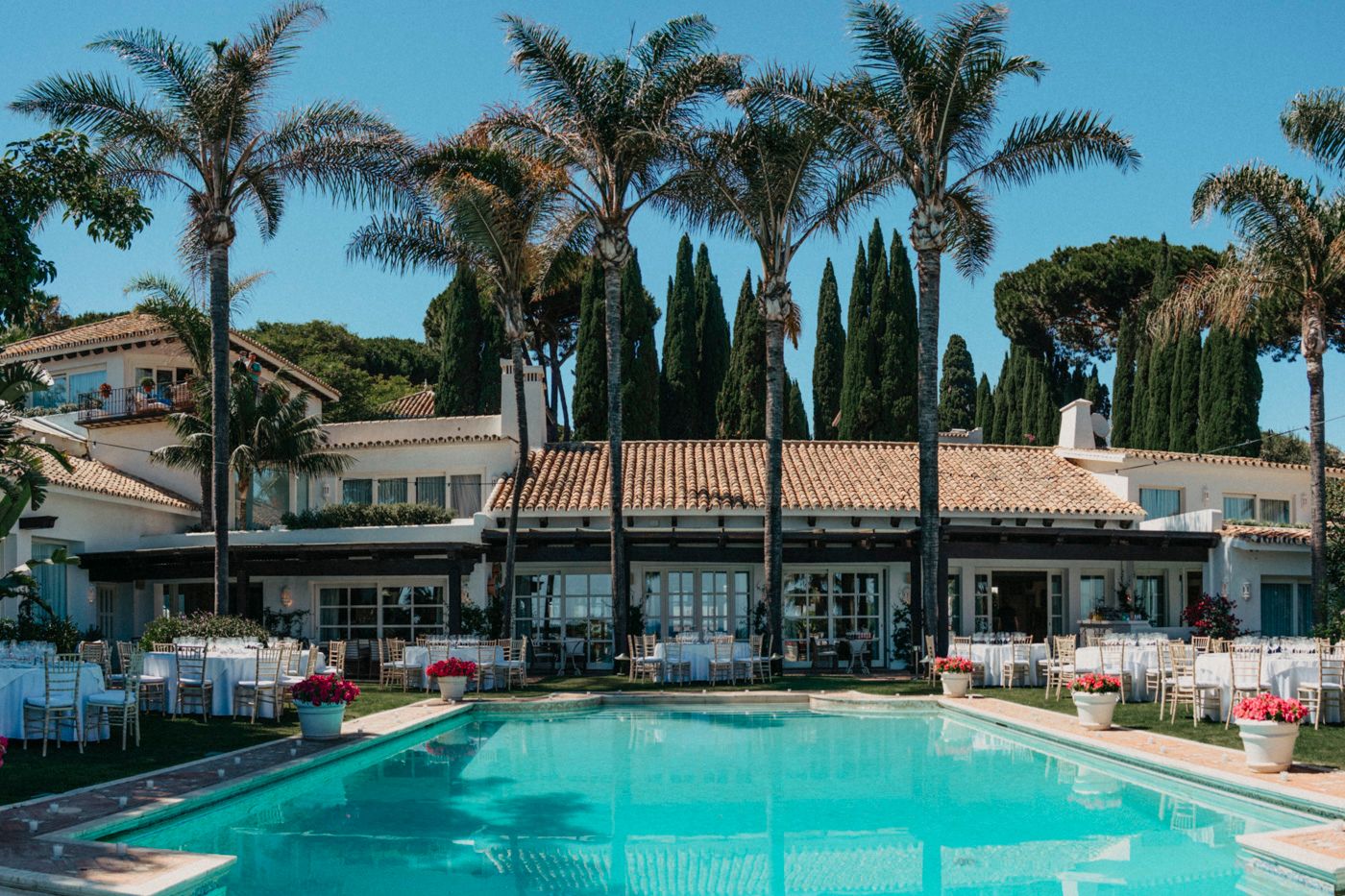 A luxury Mediterranean resort with palm trees and cypress trees surrounding a turquoise swimming pool and outdoor seating area.