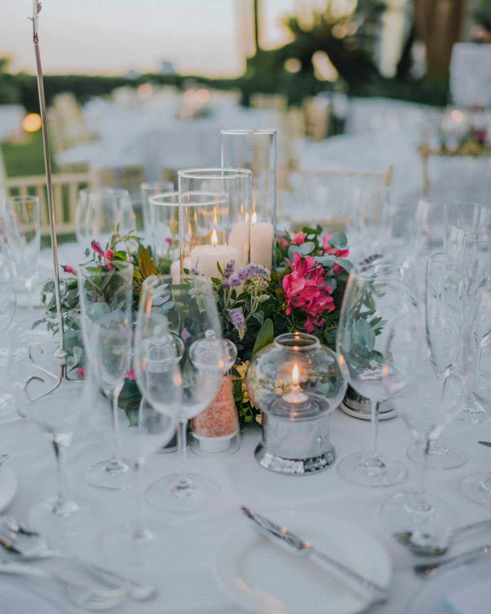 Elegant wedding table setting with pink flowers, candles in glass holders and crystal stemware on white tablecloth.