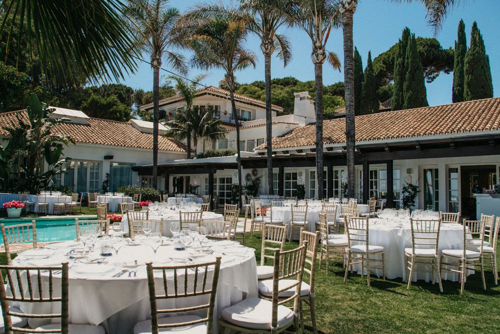 Poolside wedding reception setup with white tables and chiavari chairs surrounded by palm trees.