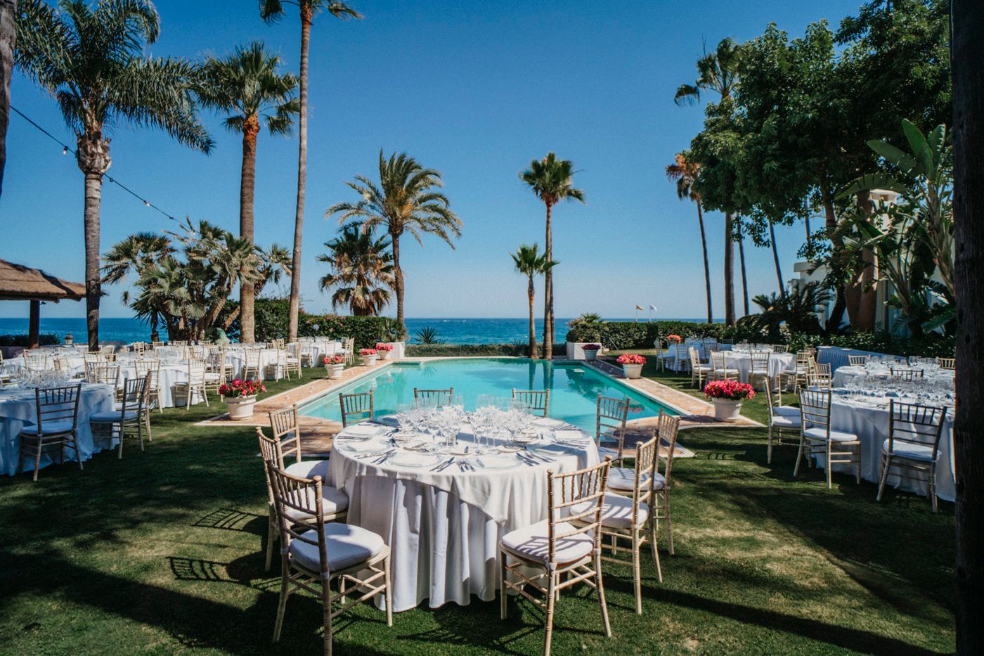 Elegant outdoor wedding reception setup with round tables near an infinity pool overlooking the ocean and palm trees.