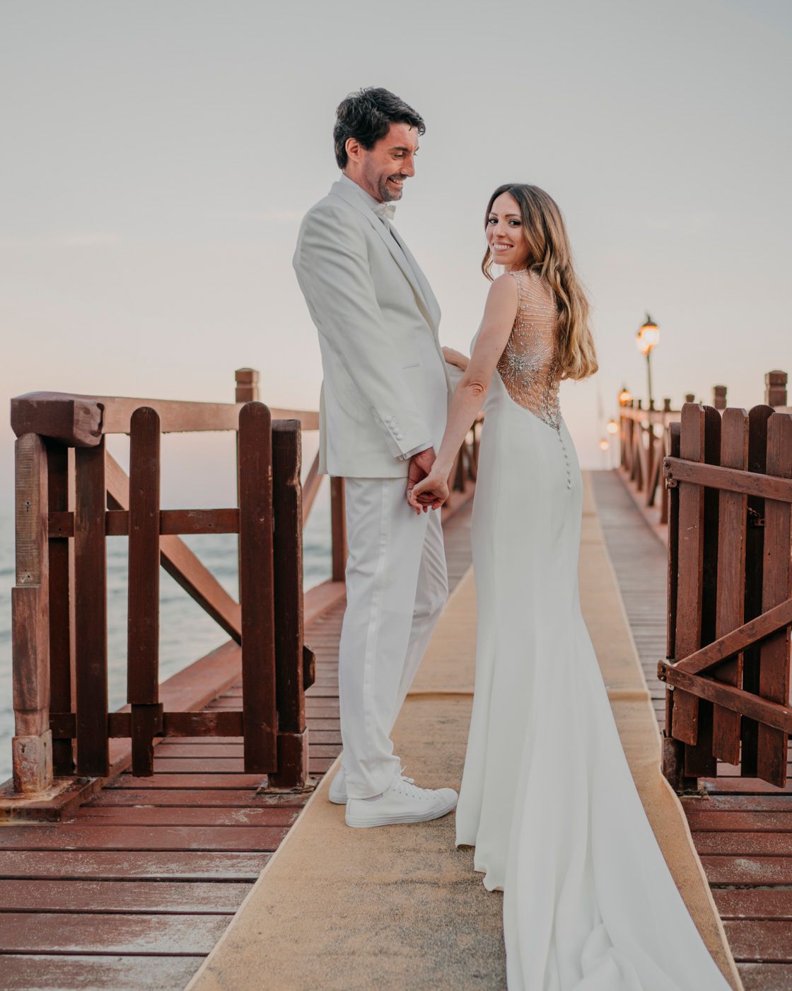 A romantic newlywed couple in white attire poses on a wooden pier at sunset.