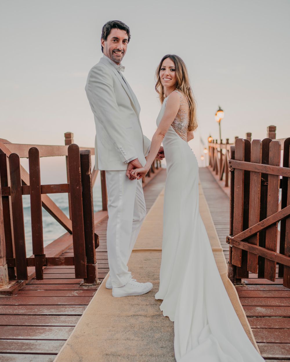 Couple in matching white wedding attire posing together on a wooden pier during sunset with a lamp post in view.