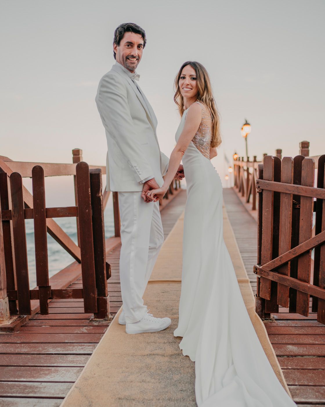 Couple in matching white wedding attire posing together on a wooden pier during sunset with a lamp post in view.