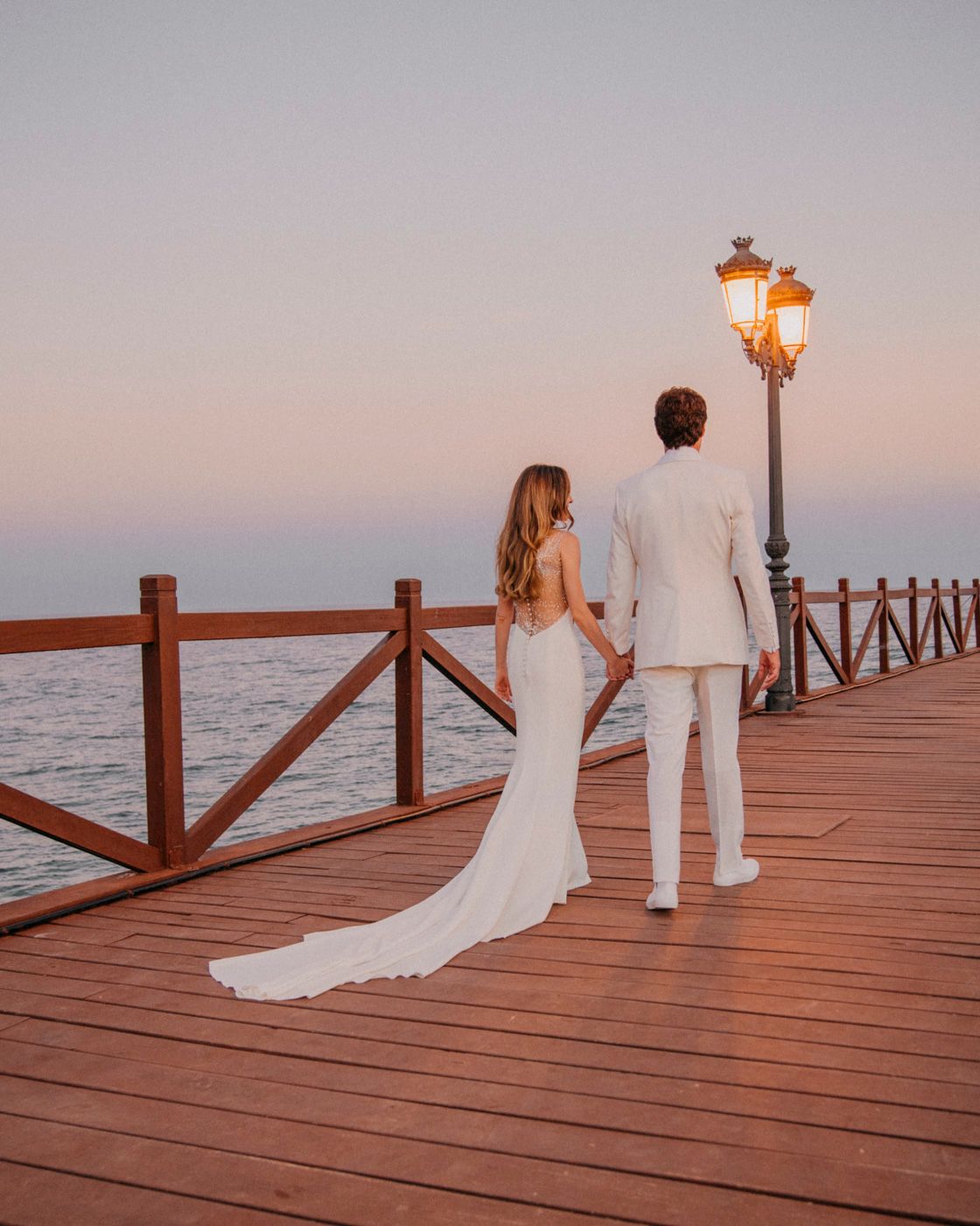 A couple in wedding attire stands on a wooden pier at sunset with a glowing vintage lamp post beside them.