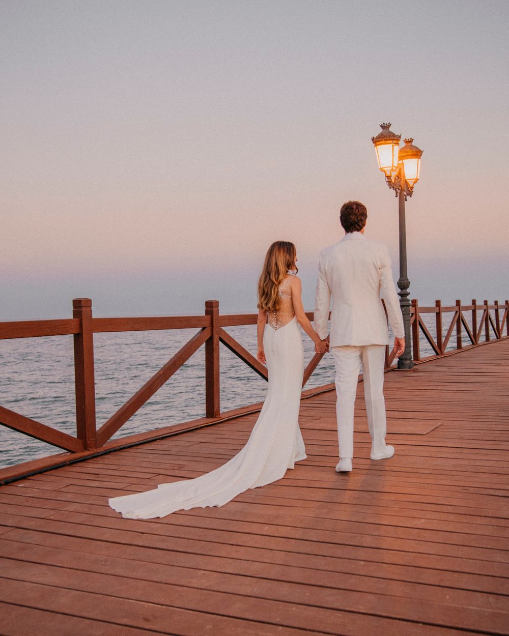 A couple in wedding attire stands on a wooden pier at sunset with a glowing vintage lamp post beside them.