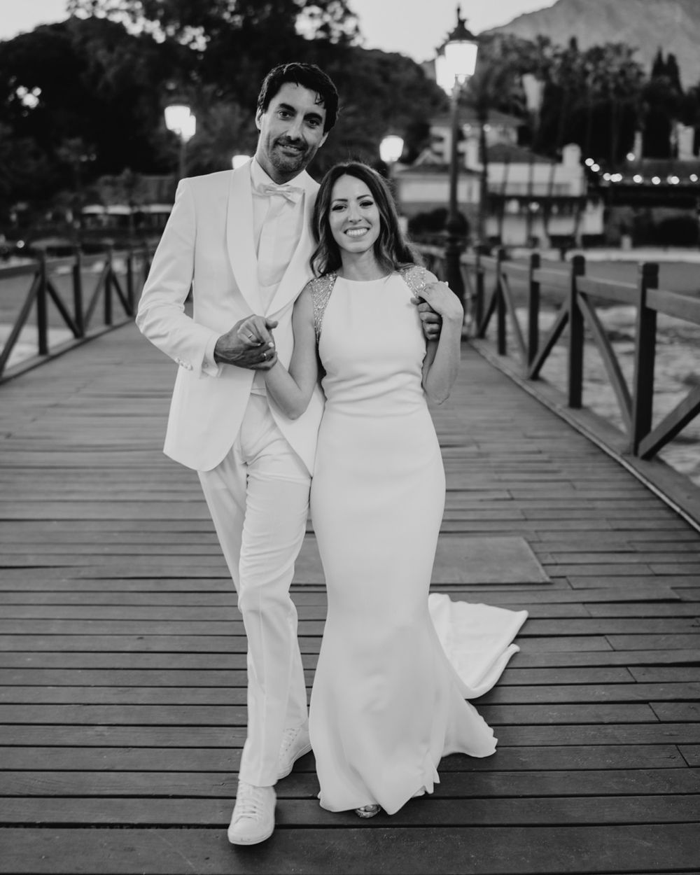 A couple in elegant white attire shares a romantic moment on a wooden boardwalk at dusk.