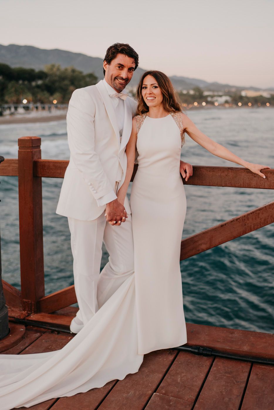 Couple in elegant white wedding attire pose together on wooden dock by ocean at sunset.