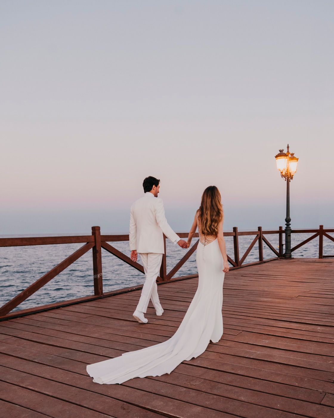 A couple in white wedding outfits standing on a wooden boardwalk by the ocean at dusk with a vintage lamp post lit.