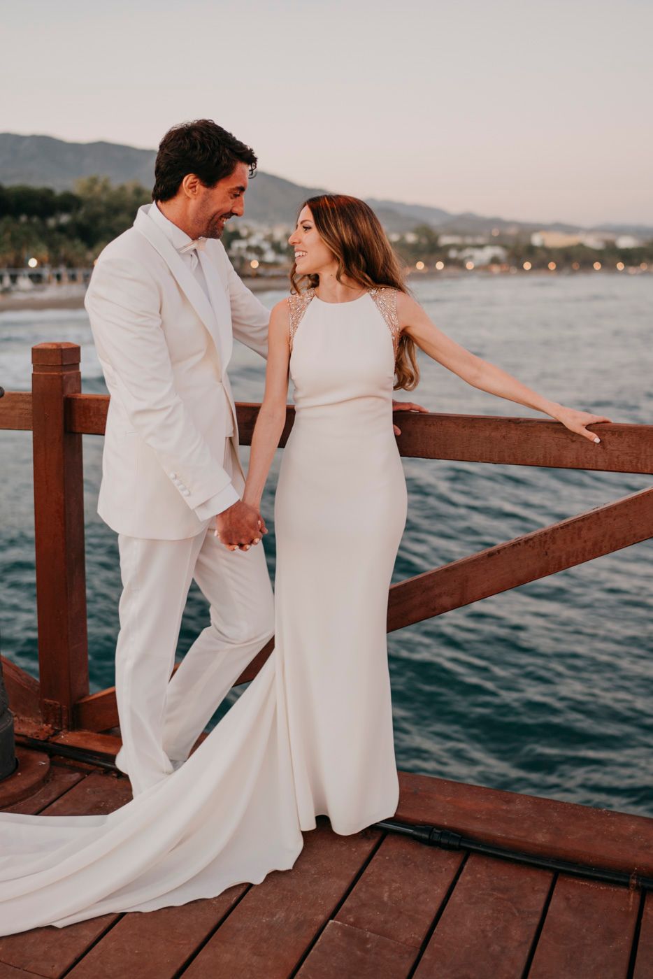 A couple in elegant white attire share a romantic moment on a wooden pier at sunset by the ocean.