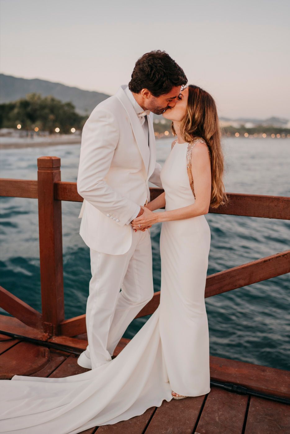 A couple shares an intimate kiss on a waterfront dock at sunset, both wearing elegant white wedding attire.
