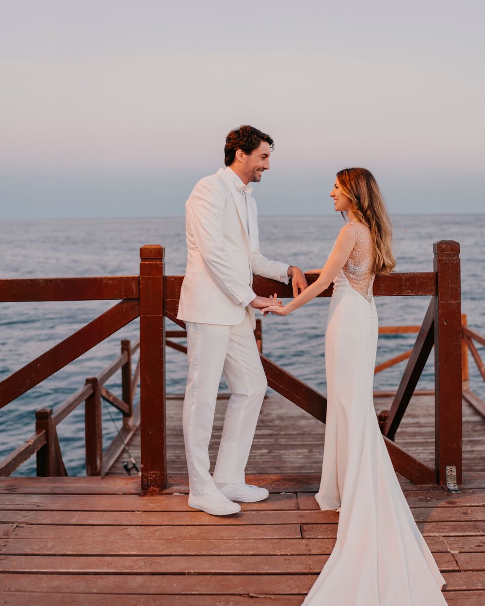 Newlyweds in matching white outfits stand on an oceanside pier during sunset.