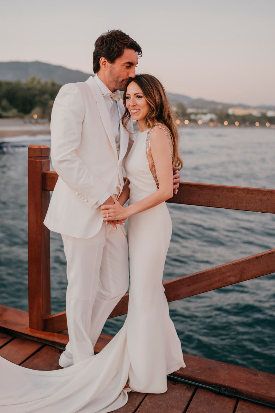 A couple in matching white formal attire embrace on a wooden dock by the water during sunset.