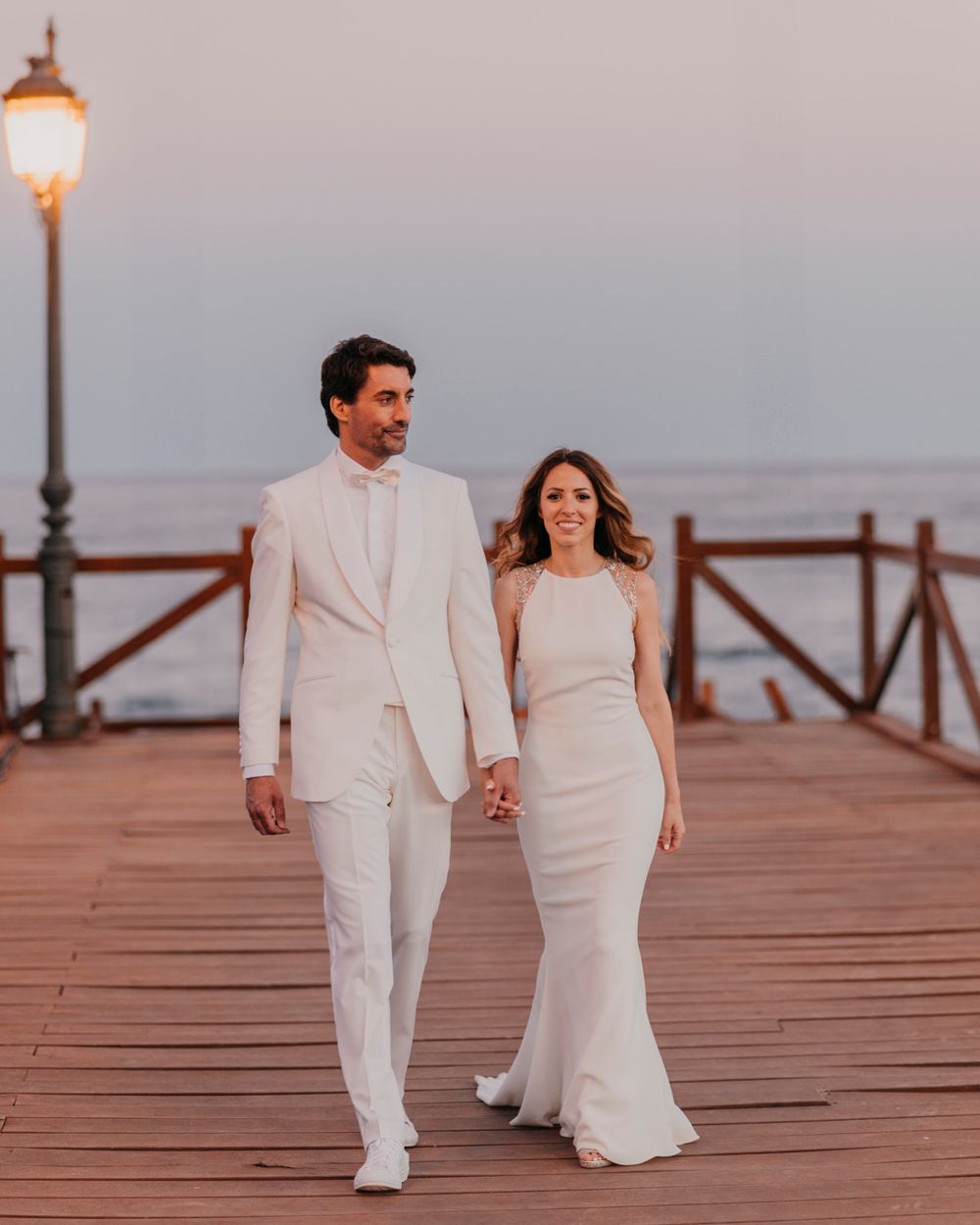 Couple in all-white wedding attire walks hand in hand along a wooden pier with a vintage lamp post.