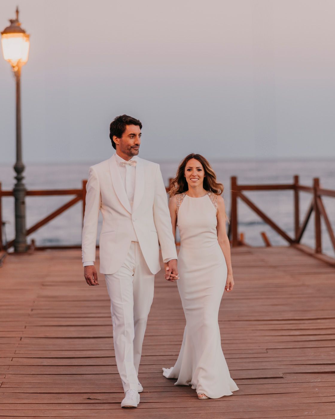Couple in all-white wedding attire walks hand in hand along a wooden pier with a vintage lamp post.