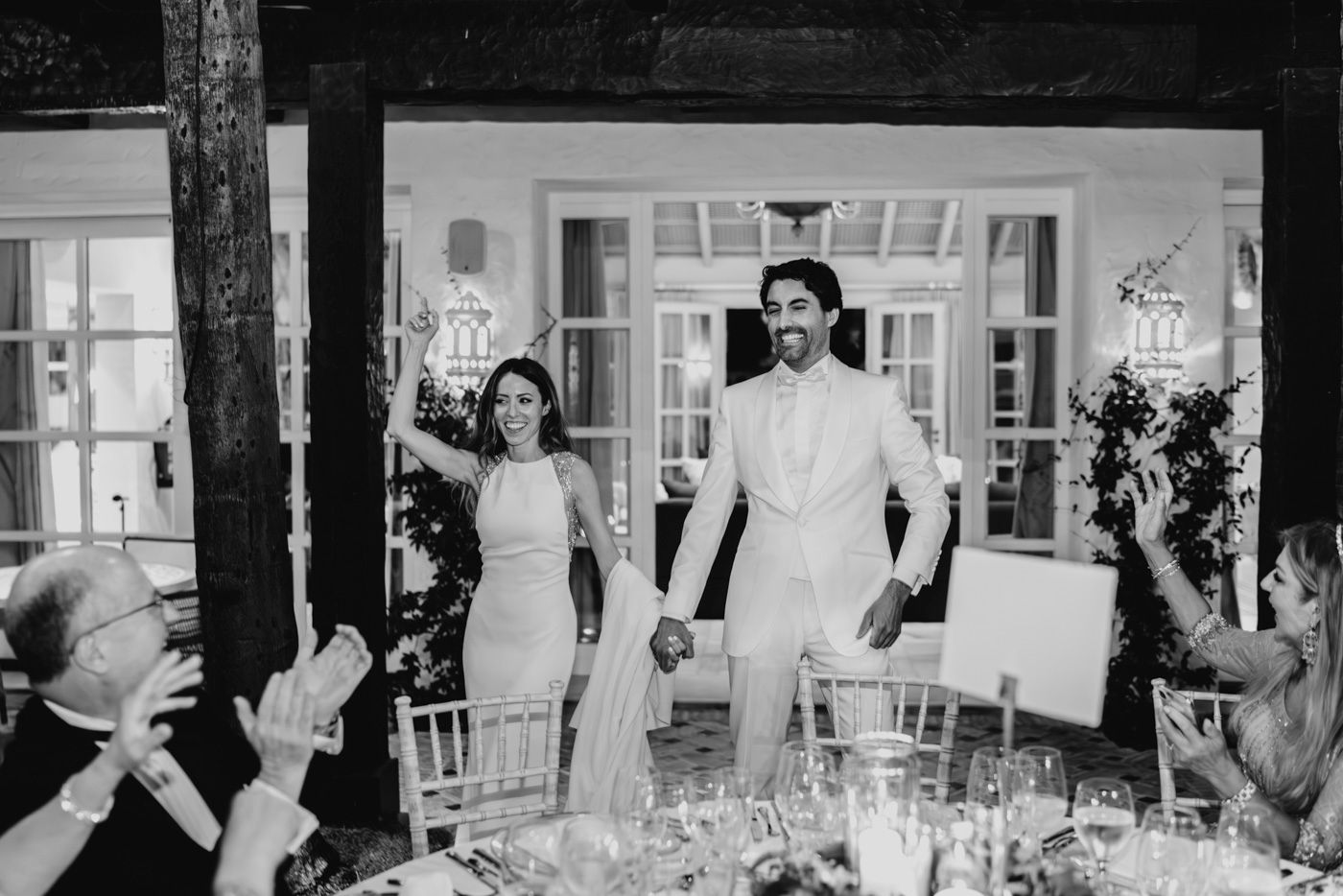 A black and white wedding reception scene with a couple celebrating among guests at an elegant table setting.