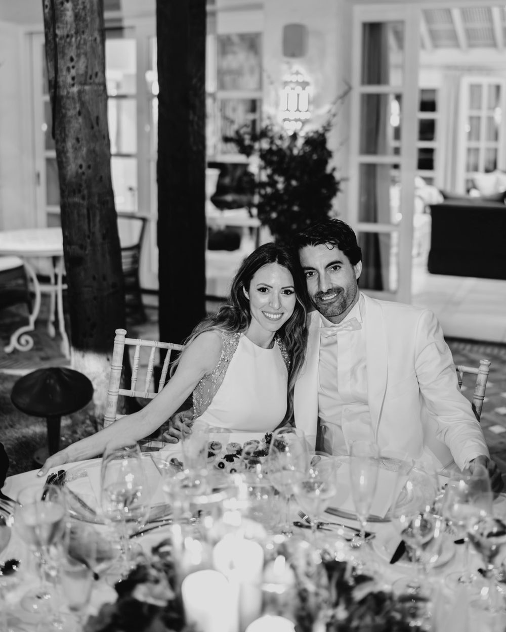 Black and white photo of couple seated together at an elegantly decorated reception table with glassware and candles.