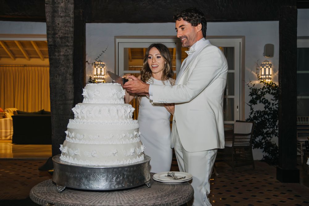 A bride and groom in light colored attire cut their elegant white tiered wedding cake together.