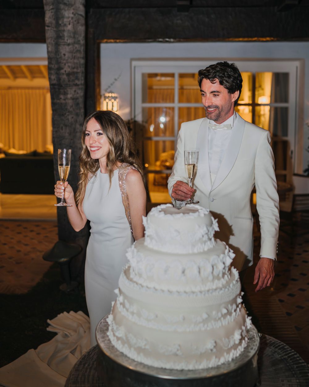 A newlywed couple poses with their elegant white tiered wedding cake and champagne glasses during their reception.