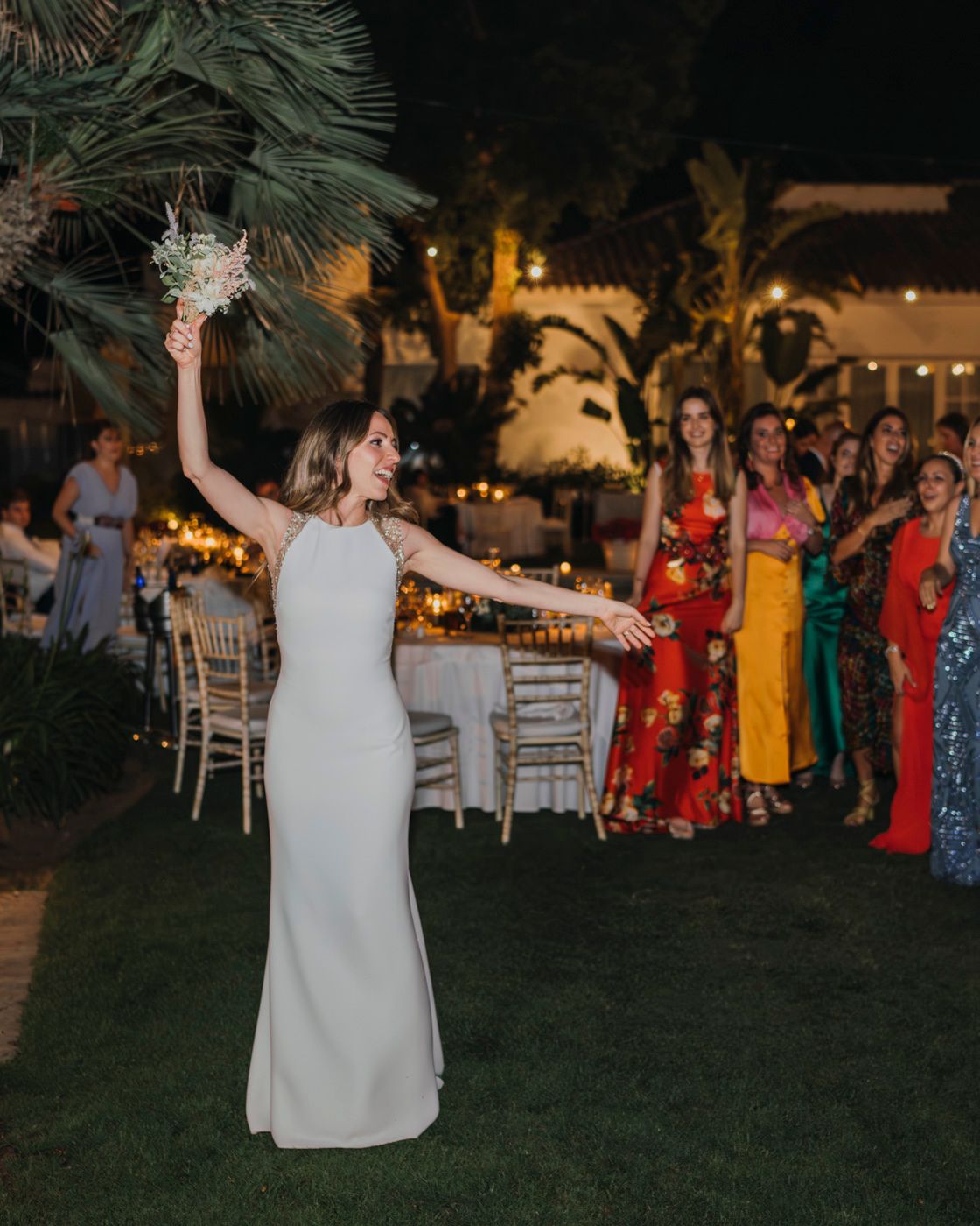 A bride in a sleek white dress celebrates while tossing her bouquet at an outdoor evening reception.