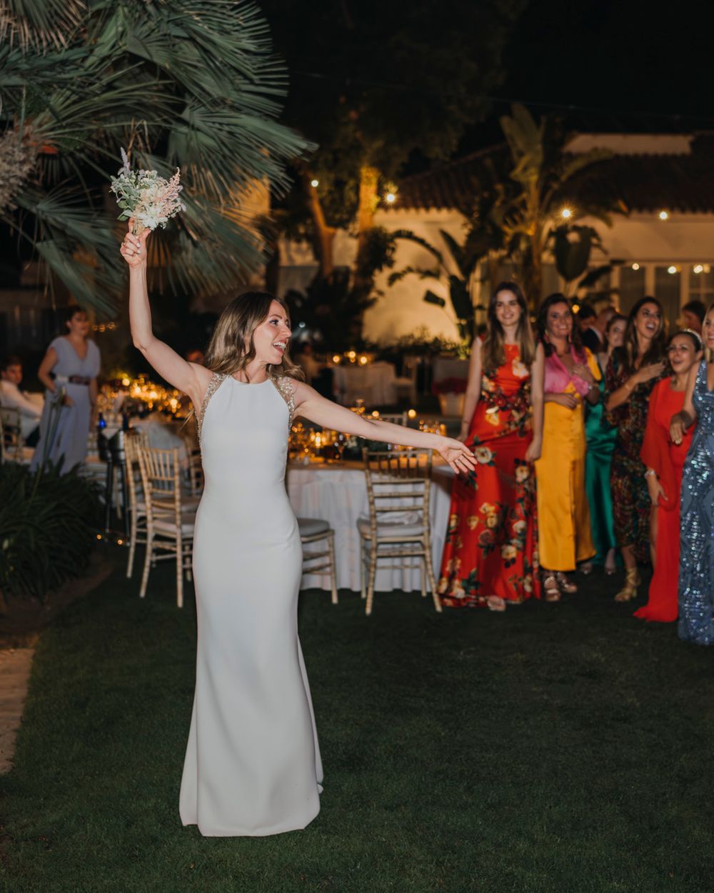 A bride in a sleek white dress celebrates while tossing her bouquet at an outdoor evening reception.