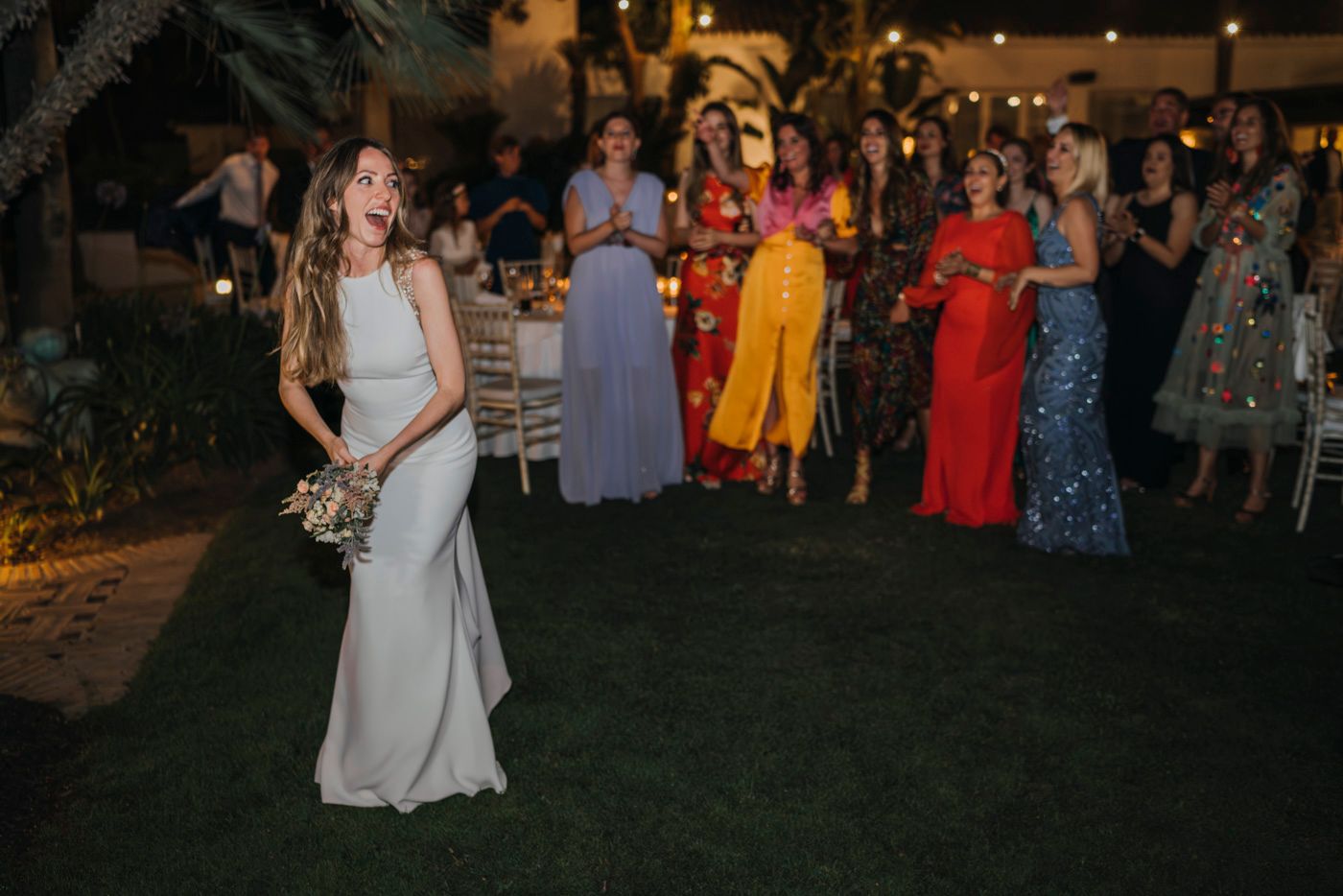 Evening wedding reception scene outdoors on grass lawn with guests in colorful formal attire watching someone in white.