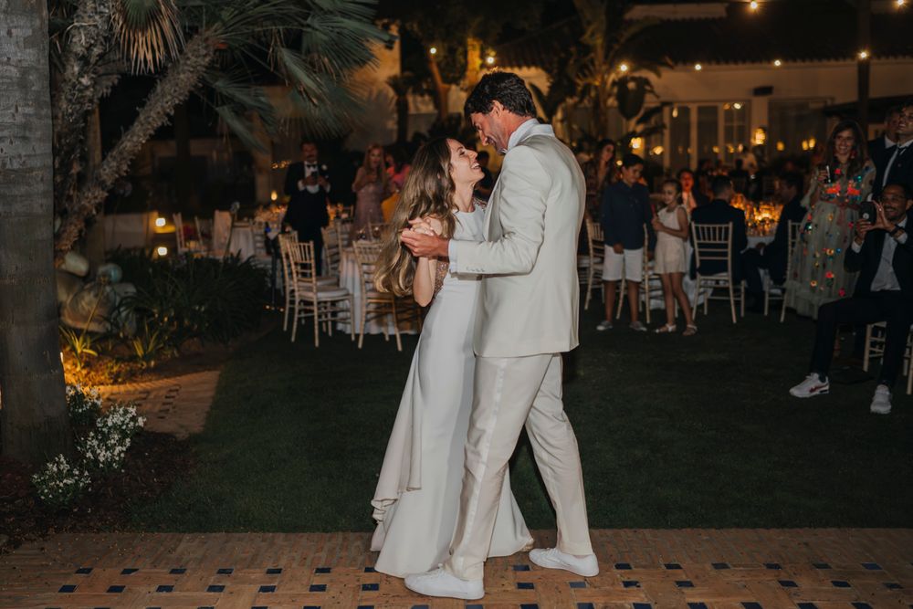 A couple shares their first dance at an outdoor evening wedding reception under string lights and palm trees.